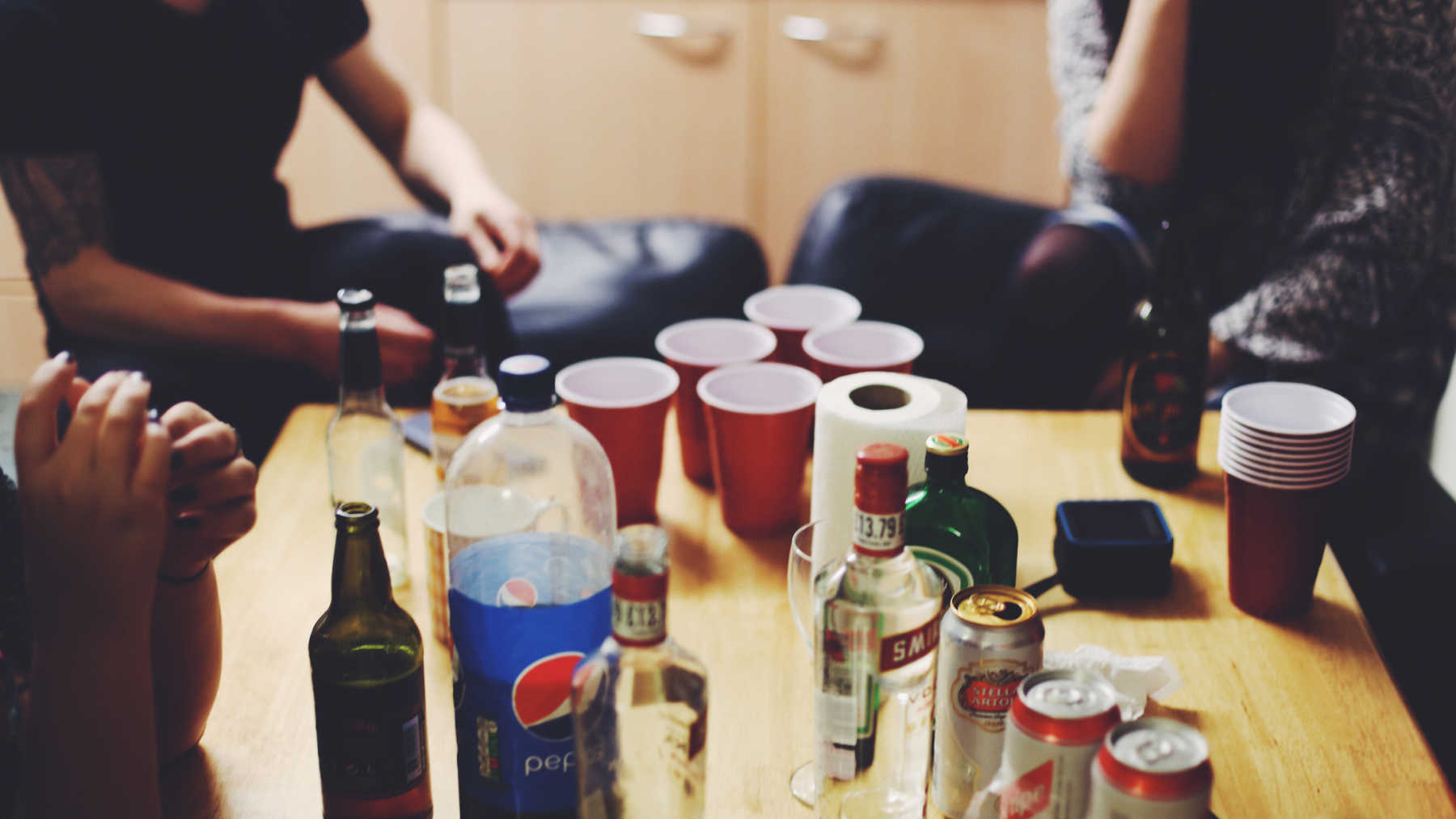 Coffee table with red solo cups, liquor bottles, open and partially consumed beer bottles with youth sitting around it.